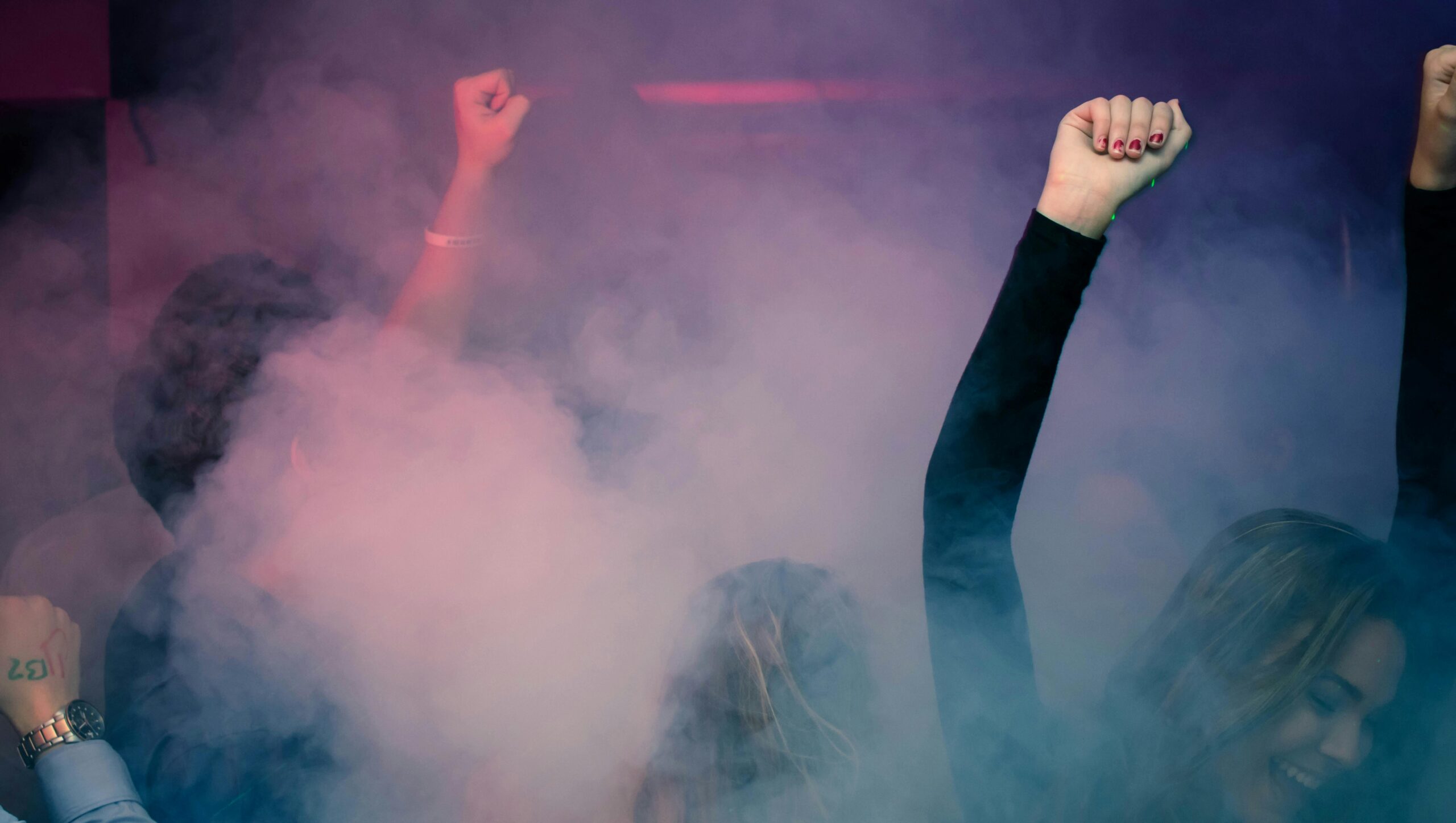 Crowd of people dancing energetically in a foggy nightclub with raised hands.