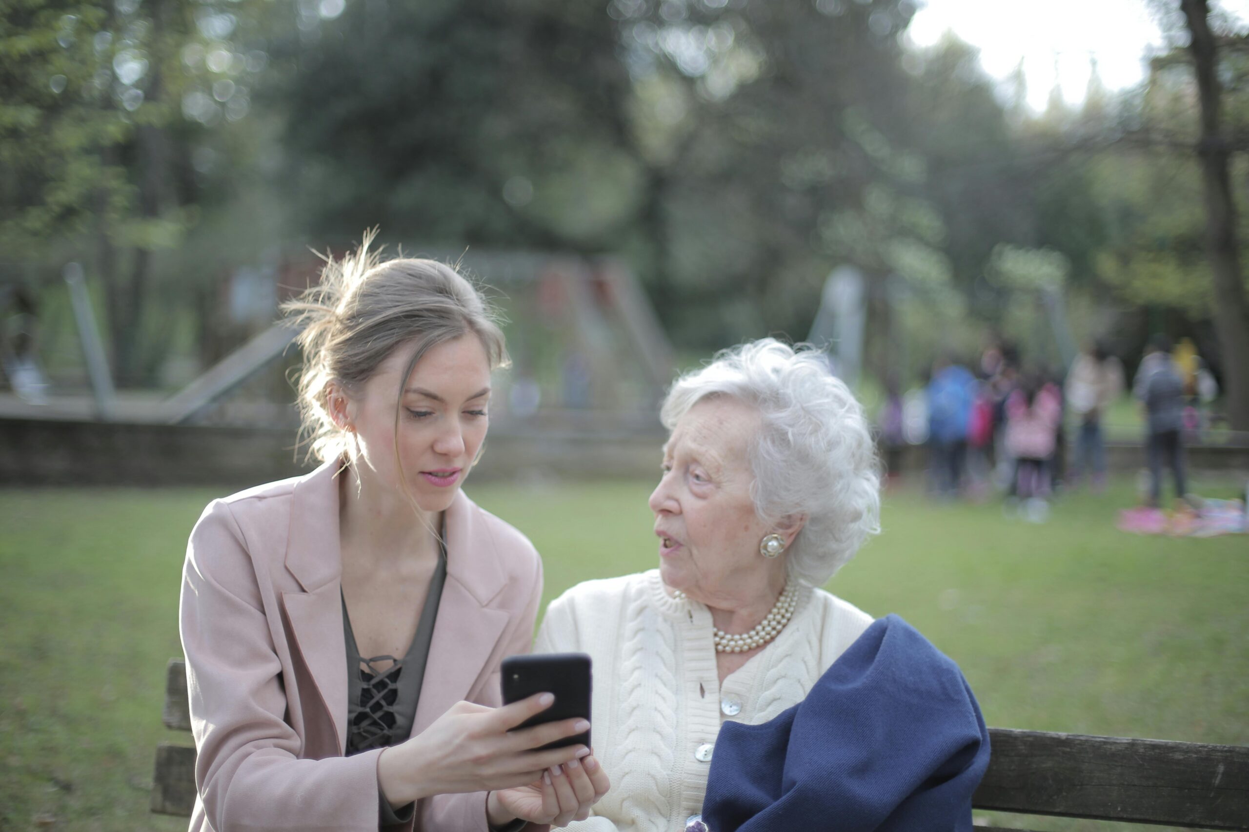 A young woman helps an elderly lady navigate a smartphone in a park, showcasing connection and support.