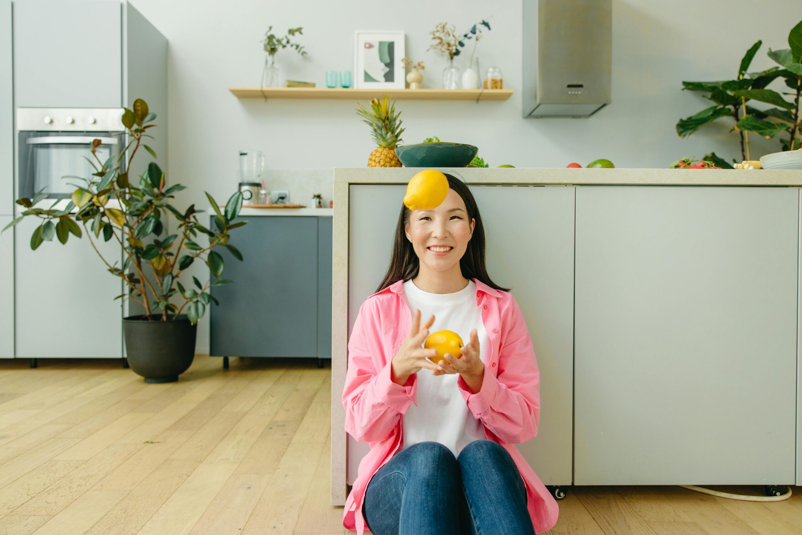 Smiling woman juggles lemons in a modern kitchen with plants and wooden flooring.